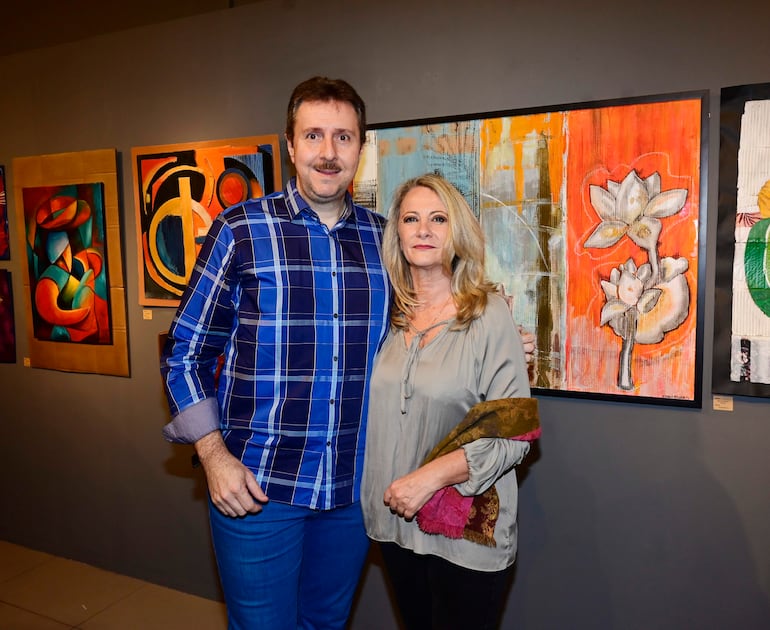 Hombre de camisa a cuadros y mujer con blusa gris sosteniendo una bufanda, ante pinturas abstractas en la galería.