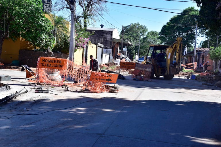 Dos trabajadores, uno agachado con abrigo oscuro, manipulando herramientas junto a una retroexcavadora en una calle de construcción.