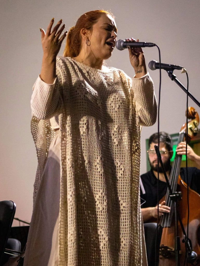 Cantante de cabello rojizo en prenda clara sostiene un micrófono y gesticula, con un contrabajista detrás en un ambiente musical íntimo.