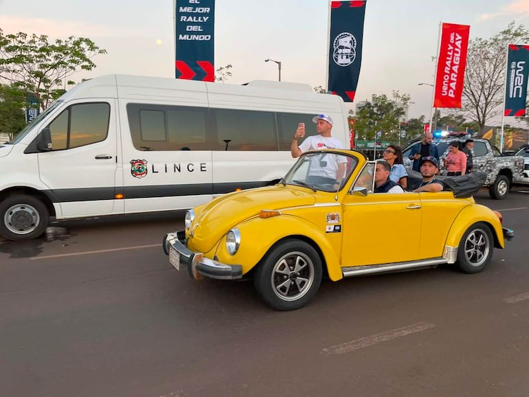 Auto convertible amarillo con tres personas, una de ellas tomando fotos. Furgoneta blanca con la palabra 'LINCE' y banderas ondeando al fondo.