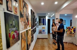 Mujer de cabello rizado escucha al hombre de mochila en galería con pinturas de cuerpos humanos y frutas.