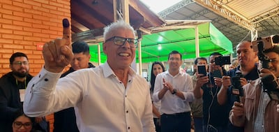 Rigoberto Chamorro sonriente, con cabello blanco y gafas, levanta el dedo marcado con tinta en un centro de votación.