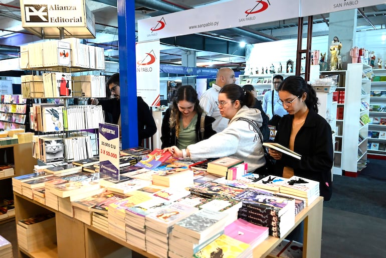 Dos mujeres revisan libros en mesa: una con sudadera clara y otra con camiseta de tirantes, rodeadas de estantes de libros.