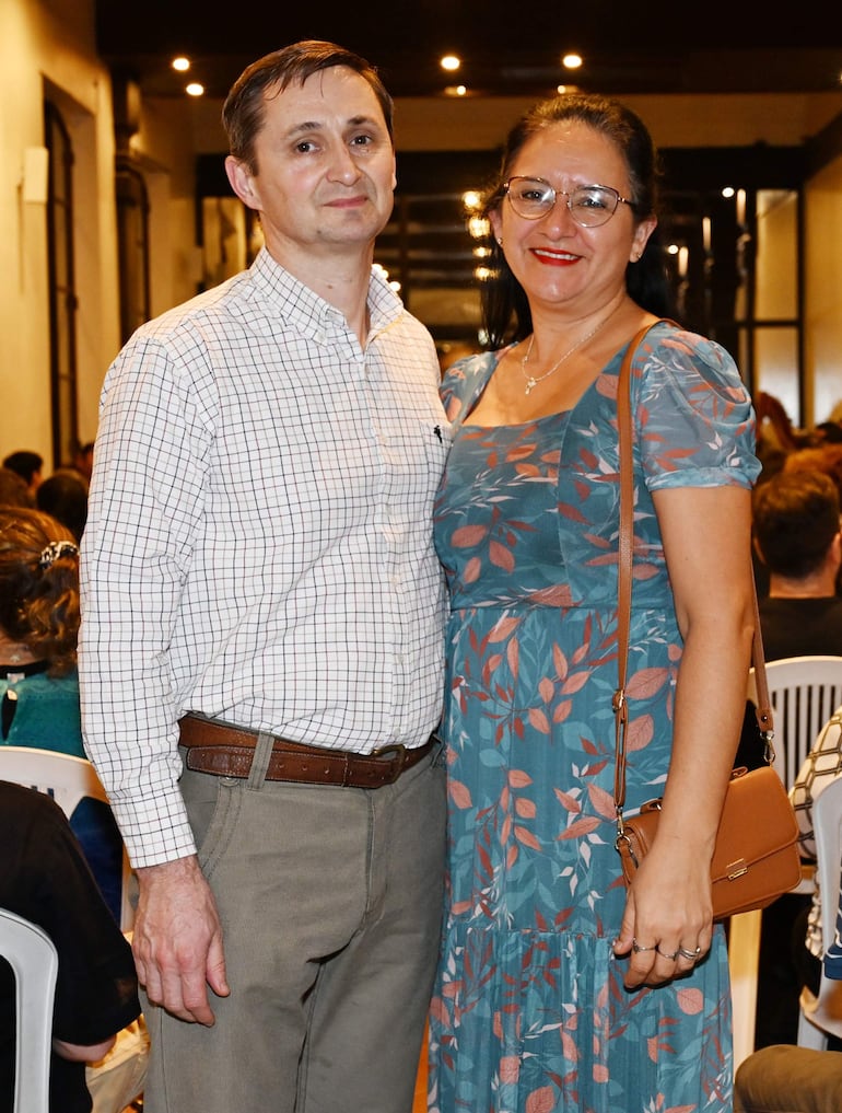 Hombre con camisa de cuadros y mujer en vestido azul floral sonríen en un salón cálido y festivo, rodeados de un público.