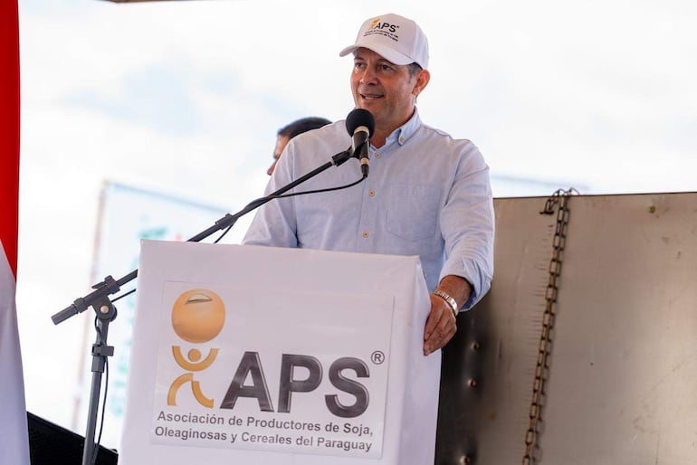Carlos Giménez en podio con camisa clara y gorra blanca de APS, hablando ante una multitud en evento al aire libre.