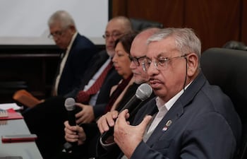 Isaías Ricardo Fretes, canoso y con gafas, habla sosteniendo un micrófono. Otros miembros escuchan atentamente en un ambiente formal.