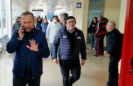 Derlis León con chaqueta azul gesticula al teléfono, rodeado de personal médico y otros en un pasillo iluminado.