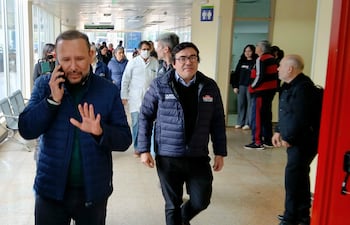 Derlis León con chaqueta azul gesticula al teléfono, rodeado de personal médico y otros en un pasillo iluminado.