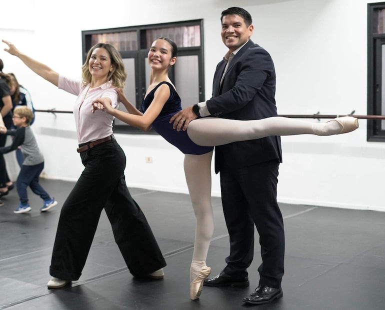 Niña sonriente en tutú negro y medias blancas, apoyada por un hombre de traje en un estudio de danza con otras personas practicando.