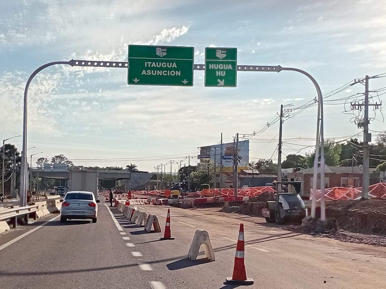 Carretera en construcción con un coche blanco en movimiento y señales hacia Itaguá y Hugua, en un ambiente urbano.