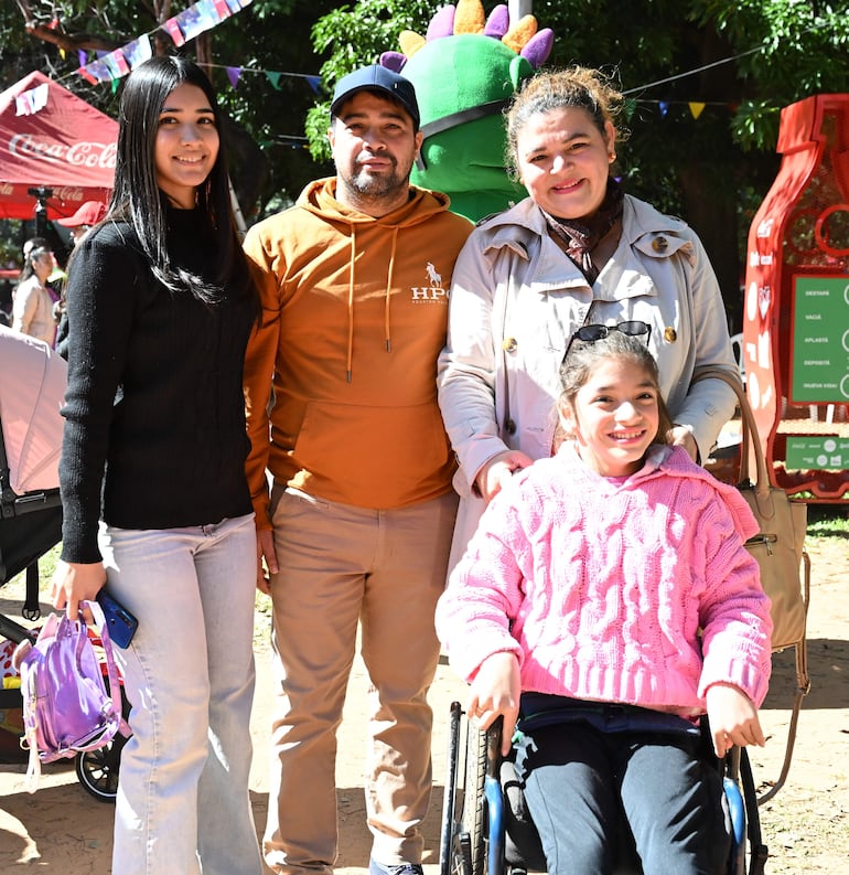 Mujer con suéter negro y jeans claros, hombre con sudadera naranja, mujer con gabardina clara y niña sonriente en silla de ruedas.