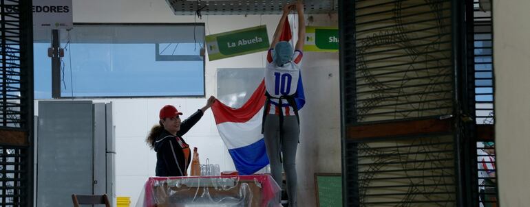 Mujeres decoran con bandera paraguaya; una lleva camiseta blanca con detalles rojos y azules, la otra viste ropa oscura y gorro rojo.