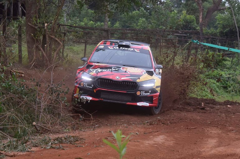 Vehículo de rally decorado con logo de Shell V-Power acelera sobre un camino de tierra, rodeado de vegetación.