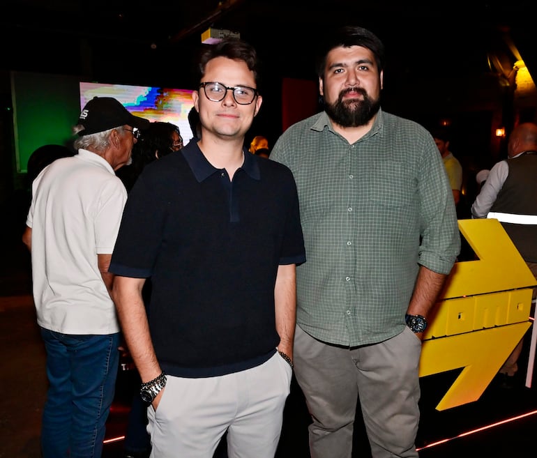 Hombre con camiseta negra y gafas sonríe junto a otro con camisa de cuadros verdes, en un evento festivo con iluminación tenue.