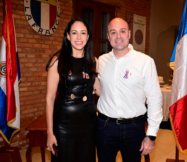 Mujer con vestido negro y distinción, y hombre con camisa blanca, posan en evento formal con banderas de Paraguay y Francia al fondo.