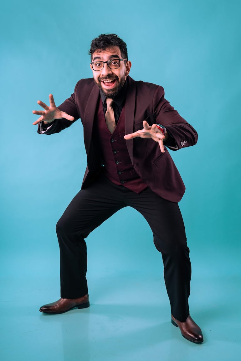 Hombre con cabello rizado y gafas, vestido con traje marrón oscuro, sonríe mientras gesticula frente a un fondo azul claro.
