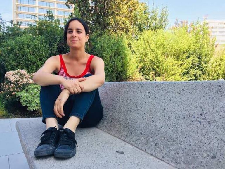 Mujer sentada en banco, camiseta de rayas rojas y negras, expresión relajada y cabello castaño suelto, rodeada de vegetación verde.