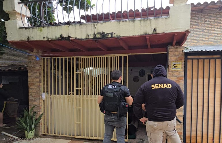 Dos agentes de la Senad en chaquetas negras monitorean frente a una vivienda con rejas en un ambiente urbano.