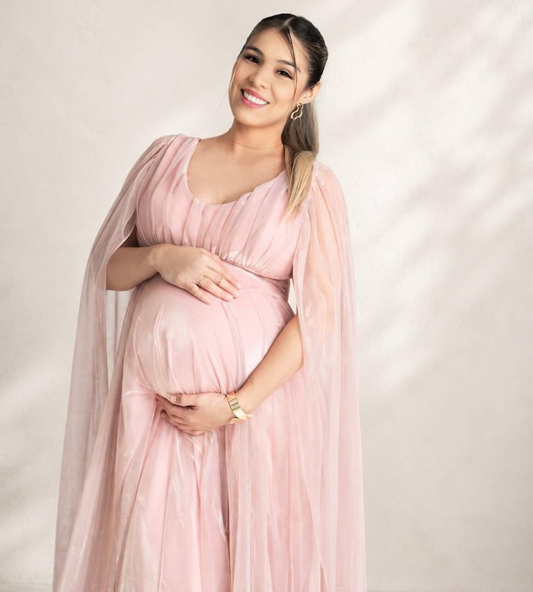 Mujer embarazada sonriente en vestido largo rosa, sosteniendo su abdomen con ambas manos en un ambiente de estudio.