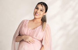 Mujer embarazada sonriente en vestido largo rosa, sosteniendo su abdomen con ambas manos en un ambiente de estudio.
