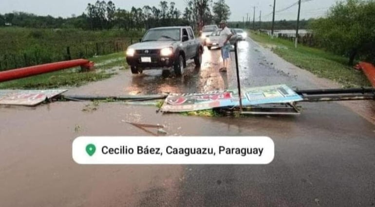 Persona observando inundación en carretera, vehículo gris detenido a un lado, ambiente lluvioso y nublado en Cecilio Báez.