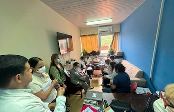 Carlos Mendoza con corbata roja en reunión con médicos, quienes toman notas en un ambiente iluminado y decorado con una pared azul.