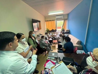 Carlos Mendoza con corbata roja en reunión con médicos, quienes toman notas en un ambiente iluminado y decorado con una pared azul.