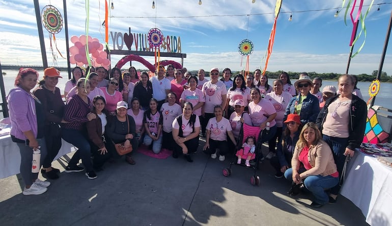 Cuarenta mujeres en camisetas rosas posan alegremente en un evento decorado con globos y cintas en un ambiente festivo junto al río.