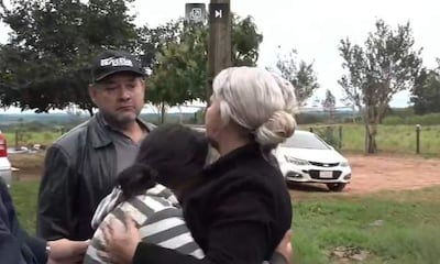 Mujeres abrazadas en lamento y hombre serio en un entorno natural, rodeados de árboles y un vehículo estacionado.