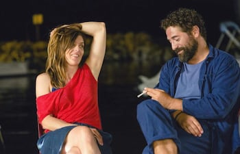 Mujer en blusa roja sonriente, reclinada, junto a hombre con camiseta y chaqueta azul, en conversación distendida al aire libre.