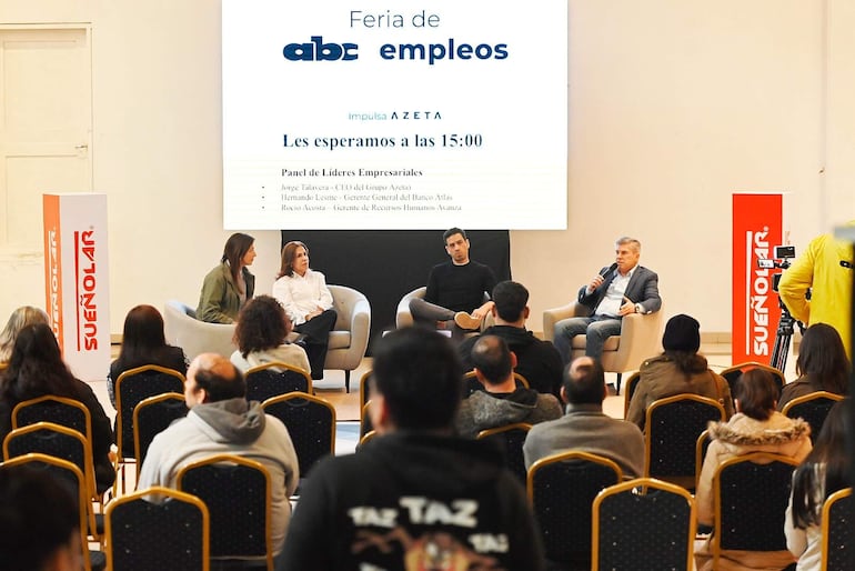 Hombre canoso habla al micrófono mientras panelistas, en atuendos formales, lo escuchan atentos en un evento de empleo.