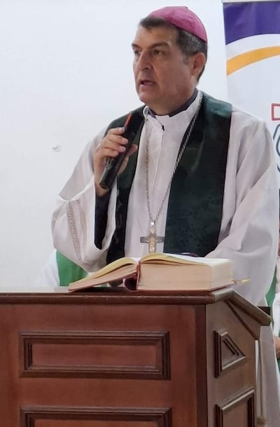 Monseñor Gabriel Escobar en vestimenta litúrgica blanca y estola verde, sostenido un micrófono con expresión seria en un ambiente religioso interior.