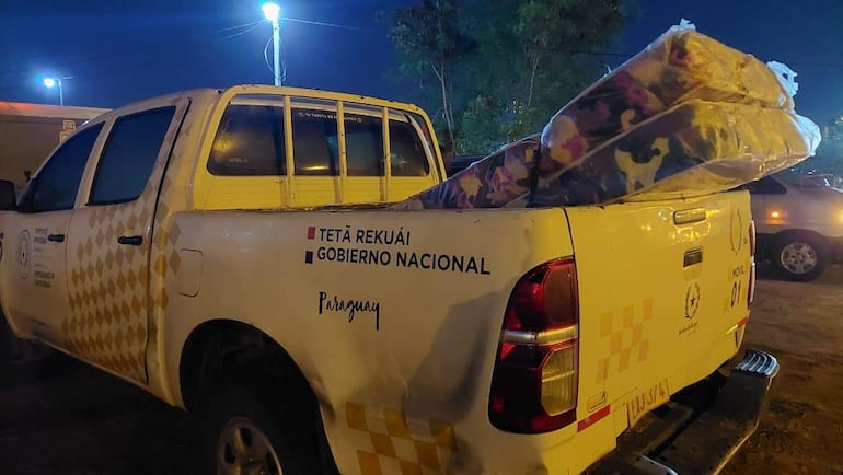Camioneta blanca con rombos amarillos y colchones en zona iluminada, simbolizando ayuda social del Gobierno Nacional.