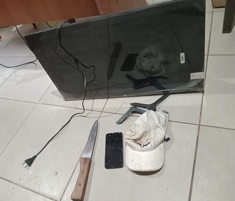Televisor JVC y cuchillo grande en el suelo, junto a un teléfono móvil y una gorra blanca sucia.