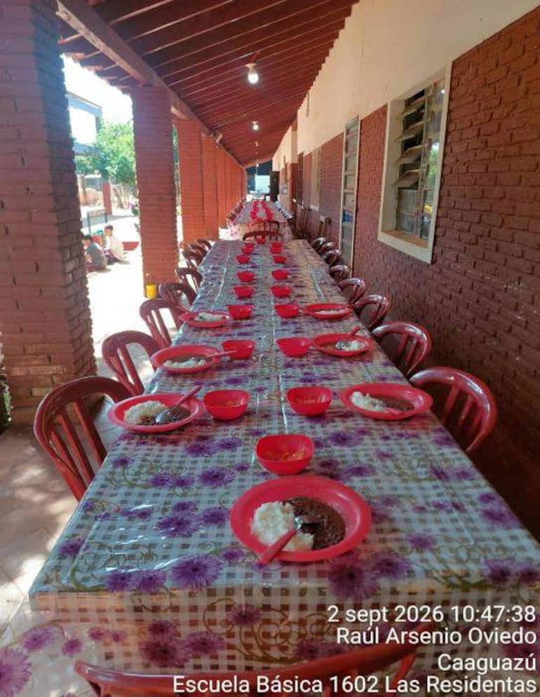 Mesa larga con mantel de cuadros y platos rojos con arroz y guiso, en un ambiente escolar sin personas visibles.