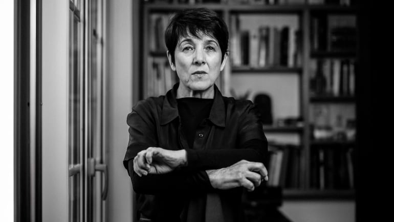 Mujer de cabello corto y oscuro, con camisa oscura, sentada con los brazos cruzados en un entorno minimalista.