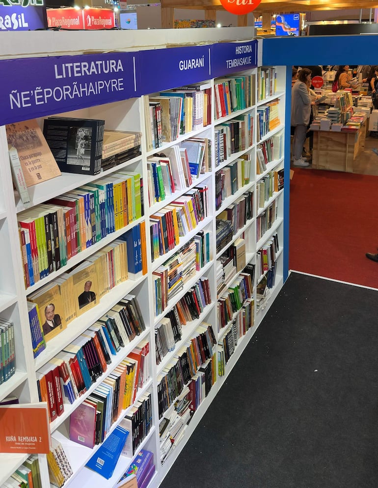 Estantería repleta de libros en Feria del Libro, con letreros de categorías y ambiente decorado. No hay personas visibles en el primer plano.