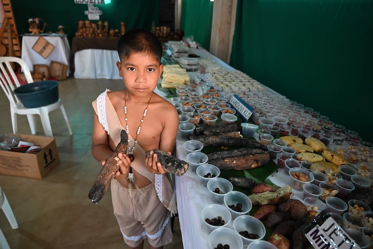Niño de la comunidad Ava Guaraní, vestido con ropa tradicional, sostiene tubérculos en un ambiente de feria con mesas de comida.