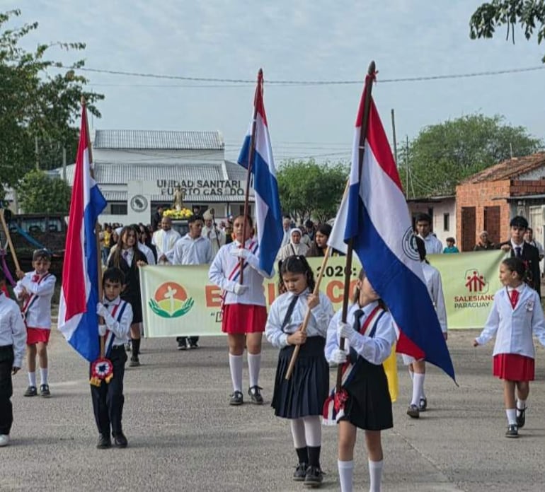 Estudiantes en uniforme blanco y azul sostienen banderas de Paraguay, mientras algunos adultos los acompañan en un ambiente festivo.