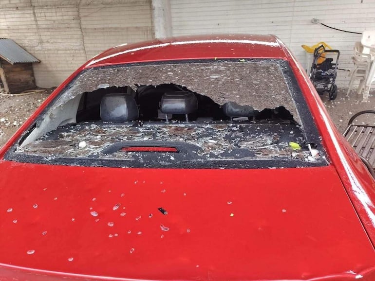 Auto rojo con techo dañado y vidrio roto, en un espacio exterior con escombros alrededor.