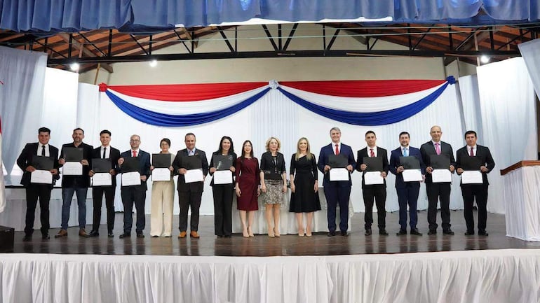 Doce personas en el escenario: seis hombres con trajes oscuros y seis mujeres en vestidos elegantes, sonriendo y sosteniendo documentos.