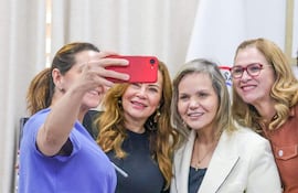Lilian Samaniego González con camiseta púrpura y Lourdes Cabrera Petters sonríen hacia la cámara en ambiente festivo con una bandera de fondo.