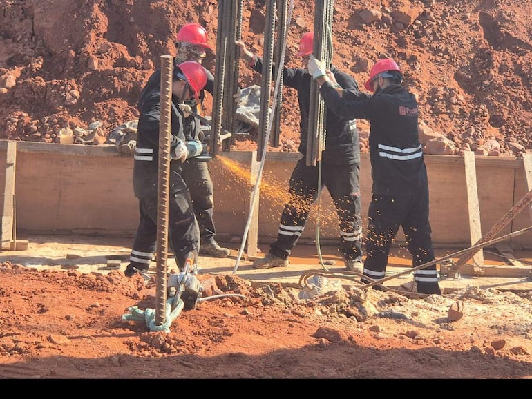 Cuatro trabajadores de construcción con cascos rojos y ropa negra sostienen barras de refuerzo en un ambiente polvoriento.
