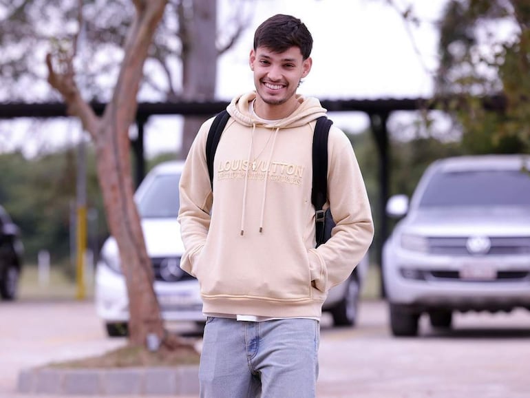 Joven con sudadera beige de Louis Vuitton y mochila negra sonríe mientras camina en un entorno urbano con coches de fondo.