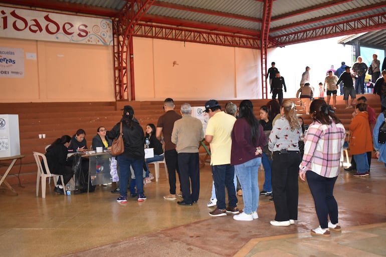 Personas en fila, algunas sentadas tras una mesa, en ambiente electoral con paredes claras y un techo de metal.