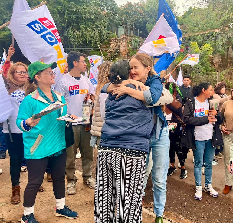 Soledad Núñez con chaqueta azul y otra mujer con chaqueta oscura se abrazan entre un grupo con banderas y folletos en ambiente comunitario.