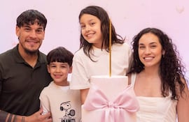Hombre con cabello rizado abraza a un niño en cumpleaños; mujer sonriente al lado, decoración rosa y torta en mesa.