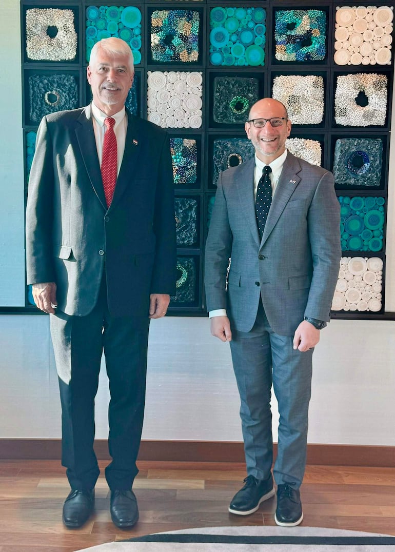 Arnoldo Wiens en traje negro y Robert Alter en traje gris claro, ambos sonriendo en un ambiente moderno con mural decorativo.