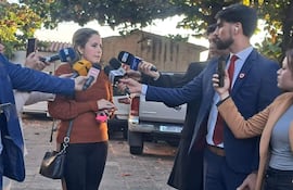Viviana Llano con suéter marrón, sosteniendo un móvil, rodeada de periodistas con micrófonos en una conferencia al aire libre.