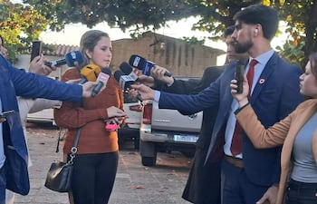 Viviana Llano con suéter marrón, sosteniendo un móvil, rodeada de periodistas con micrófonos en una conferencia al aire libre.